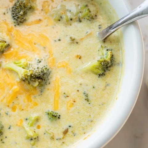 A close-up of velvety Broccoli Cheddar Soup in a rustic bread bowl, topped with fresh florets and a swirl of heavy cream.  
